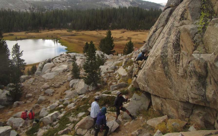 A group of people rock climb on a steep rock wall in the foreground, with an alpine meadow, lake and evergreen trees appear in the background. 